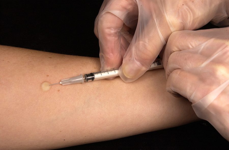 Impfung. Zwei Hände in Schutzhandschuhen setzen eine Spritze in den Unterarm.